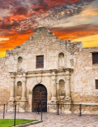 Elm Hollow Apartments stand majestically like a historic stone mission building, silhouetted against a sunset sky, with the Texas flag gently waving in the breeze.