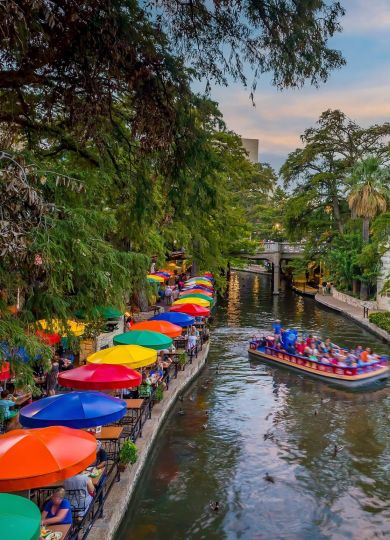 Colorful umbrellas line the vibrant San Antonio River Walk, echoing the lively community at Elm Hollow Apartments, as a boat passes under a stone bridge.